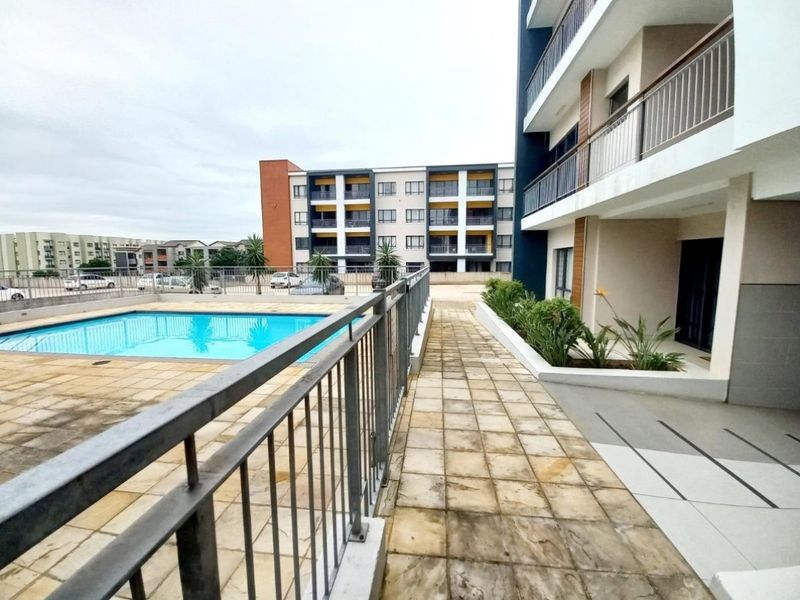 A well-maintained communal swimming pool area featuring paved decking, metal safety railings, and landscaped garden beds within an apartment complex.