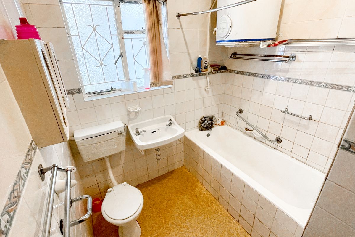 A dated bathroom featuring a standard bathtub, wall-mounted basin, and toilet, with tiled walls and a window with security bars.
