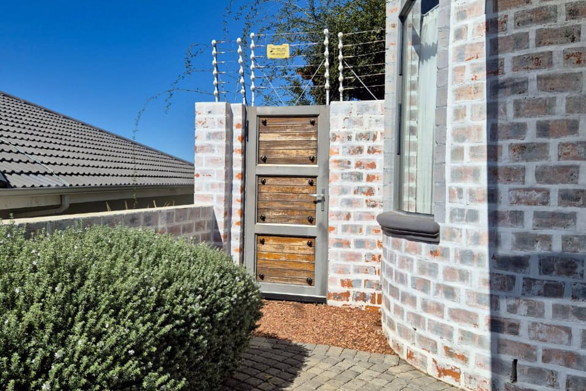 A private side entrance featuring a rustic wooden gate, brickwork facade, and paved walkway, secured with electric fencing.