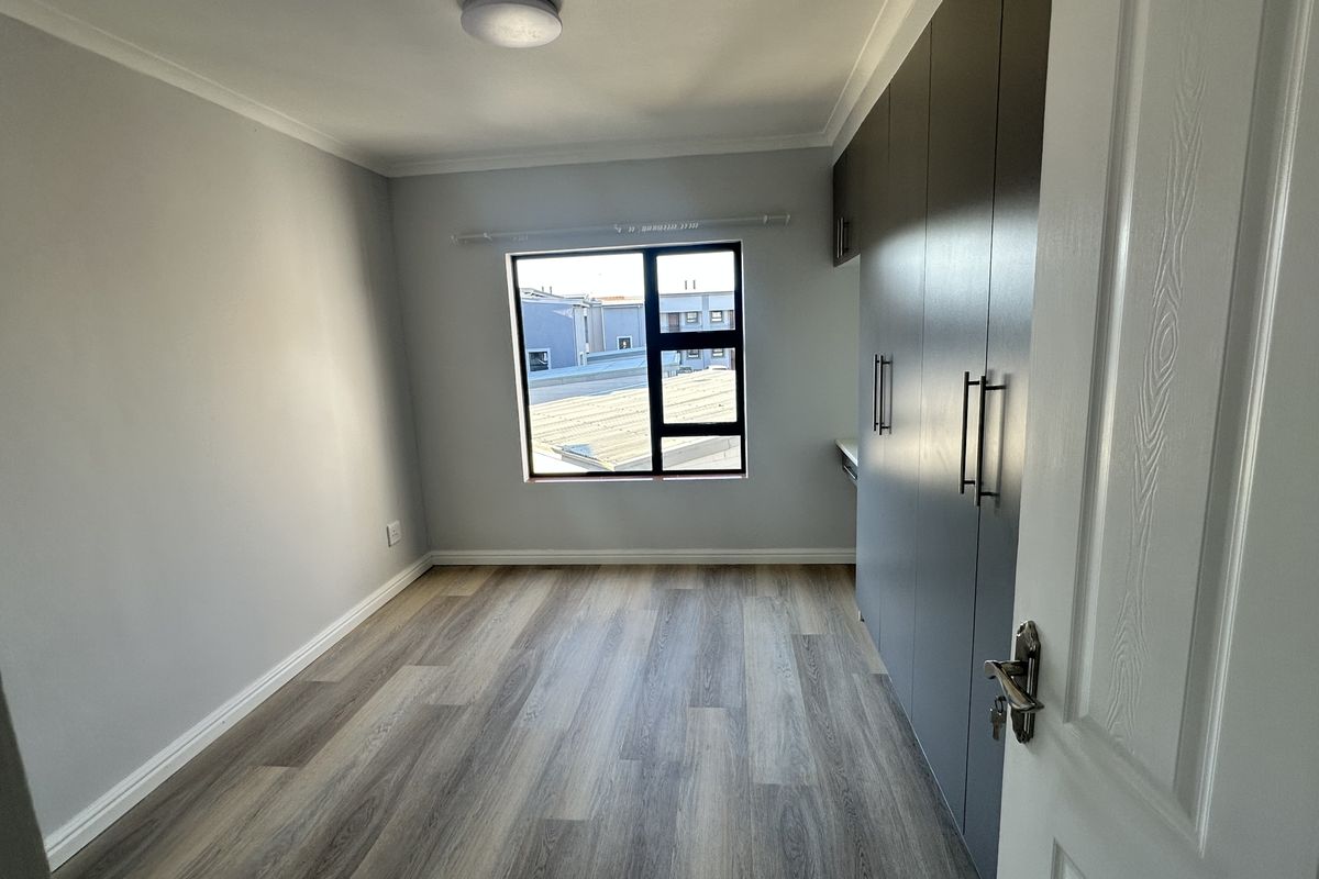 A bright, empty bedroom featuring modern wood-look laminate flooring, neutral grey walls, and extensive built-in dark grey cabinetry with integrated desk space.
