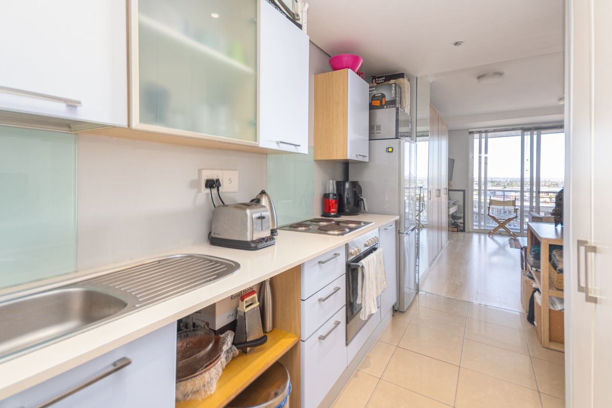 Compact galley-style kitchen featuring white cabinetry, integrated oven and hob, stainless steel sink, and tiled flooring, leading into an open-plan living area.