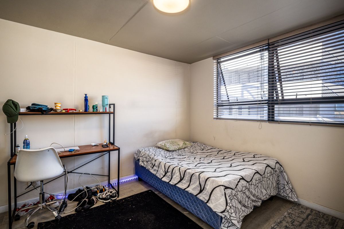 A functional bedroom featuring light-toned flooring, a large window with fitted blinds, and a neutral color palette.