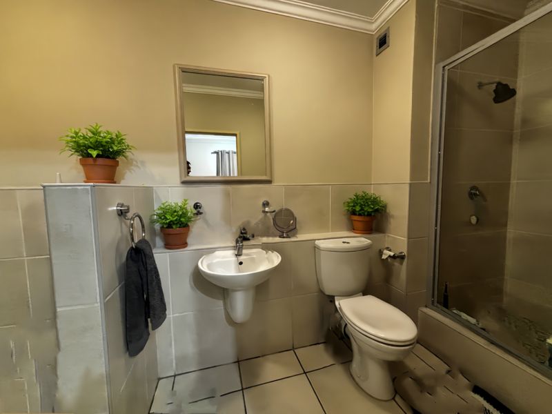 A functional bathroom featuring a pedestal basin, a standard toilet, and a glass-enclosed shower with neutral wall and floor tiling.