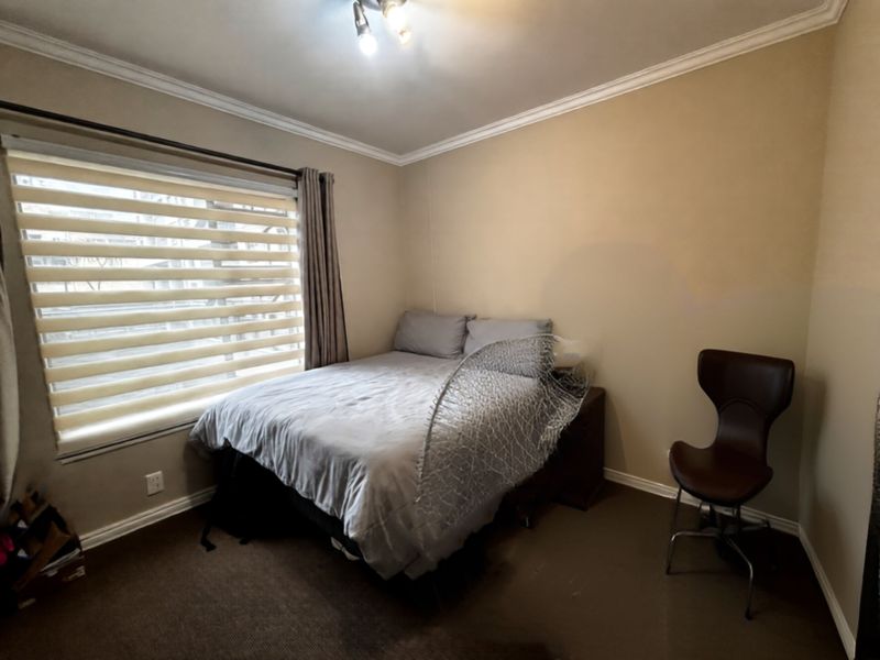 A functional bedroom featuring neutral wall tones, carpeted flooring, and a large window with fitted blinds providing natural light.