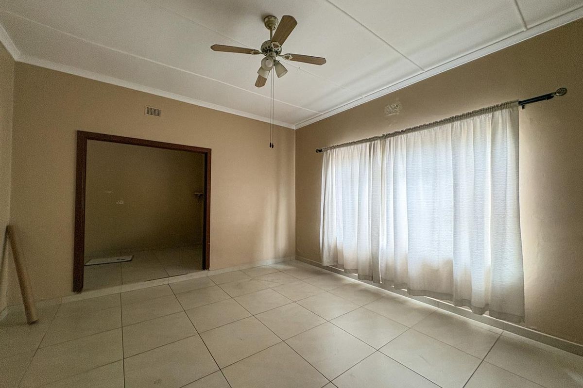 A spacious, empty living room featuring neutral tiled flooring, a ceiling fan, and large windows with curtain rods, offering a blank canvas for personalization.