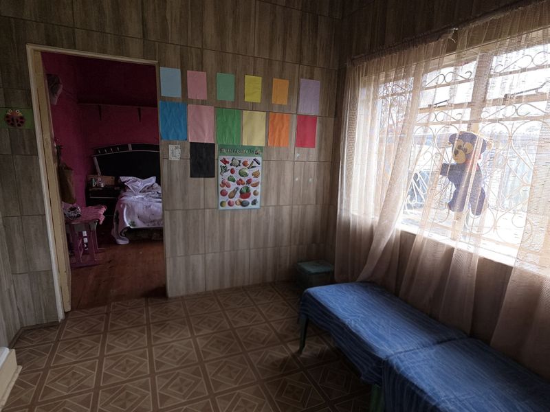 A living area featuring wood-look wall tiles, tiled flooring, and a large window with a security gate, leading into an adjacent bedroom.