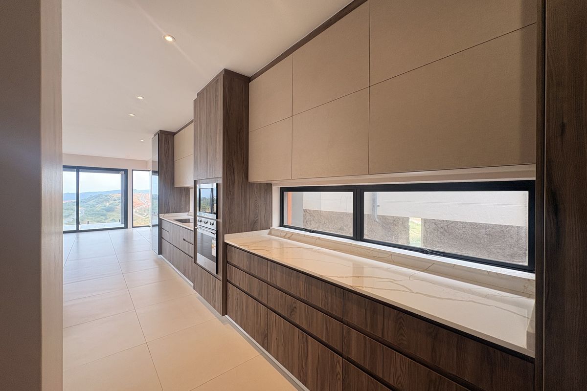 Modern kitchen featuring sleek wood-grain cabinetry, stone countertops, and integrated oven, flowing into an open-plan living area with expansive views.