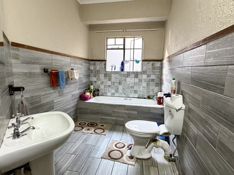 A functional bathroom featuring a bathtub, pedestal basin, and toilet with grey tiled walls and flooring, requiring some maintenance to the exposed plumbing.