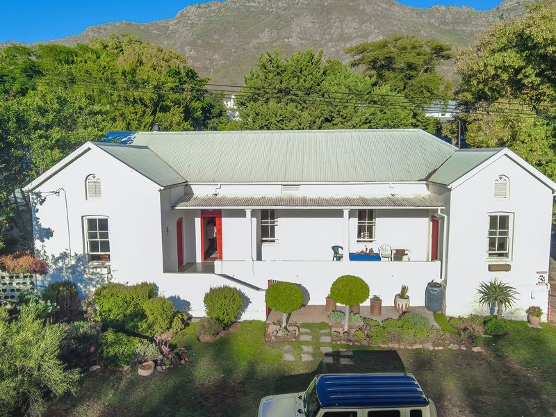 A charming single-story residence featuring a classic corrugated metal roof, a covered front veranda, and established garden surroundings set against a mountain backdrop.
