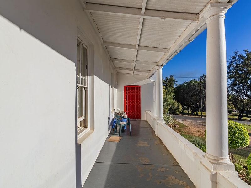 A covered veranda featuring classical columns, a red security gate, and a painted concrete floor overlooking the front garden.