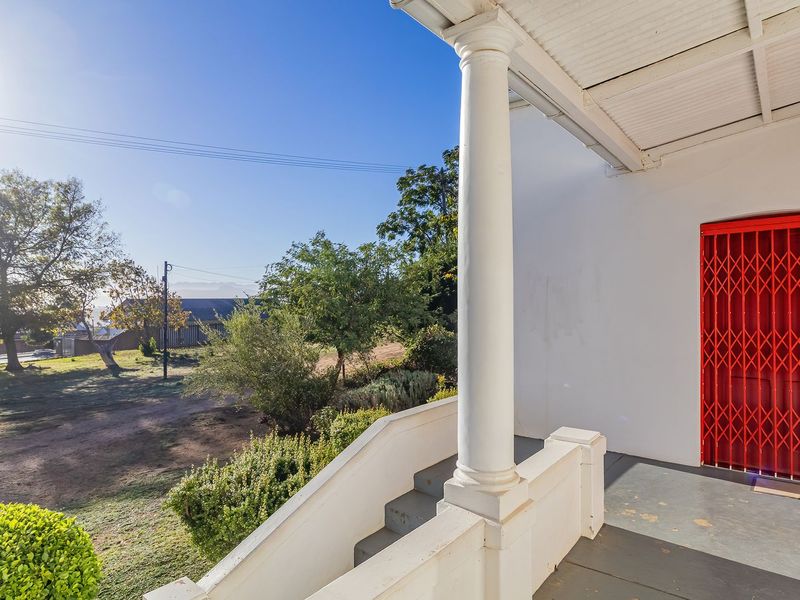 A covered veranda featuring a classical column, concrete stairs, and a bright red security gate, overlooking a spacious garden area.