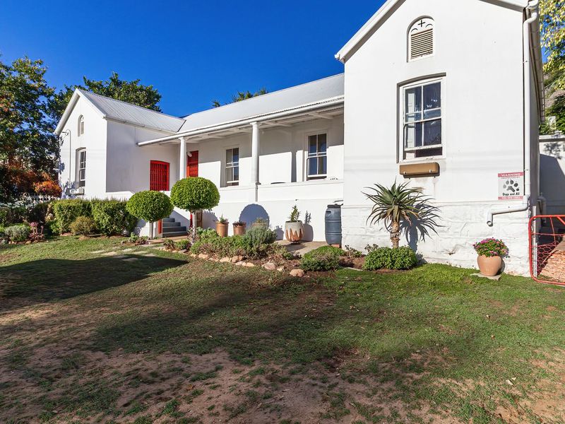 A charming white cottage-style home featuring a covered veranda, metal roofing, and classic sash windows, set within a landscaped garden.