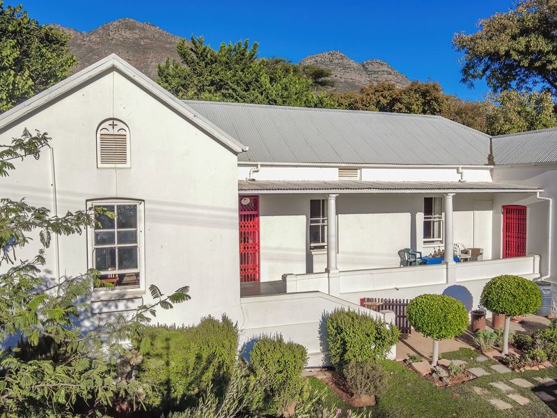 A charming white cottage-style home featuring a covered veranda, classic columns, and a corrugated metal roof, set against a scenic mountain backdrop.