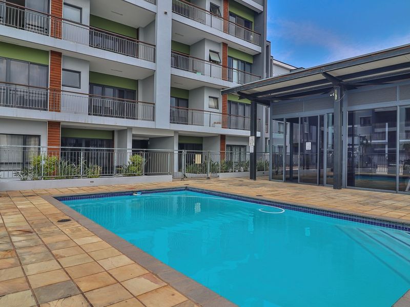 Communal swimming pool area featuring paved decking and a covered structure, set within a modern apartment complex.