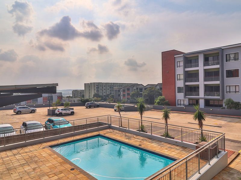 Elevated view of the apartment complex featuring a communal swimming pool, paved pool deck, metal safety railings, and adjacent parking area.
