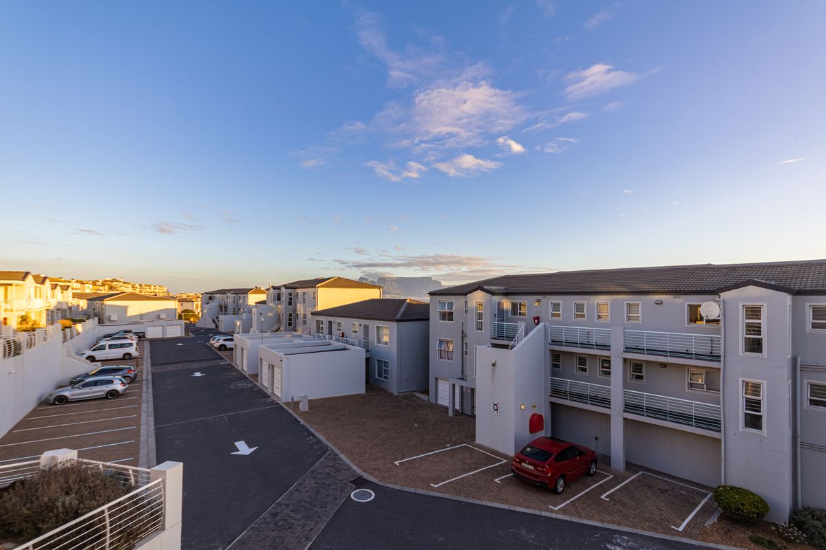 An elevated view of the apartment complex exterior, featuring paved parking areas, multi-story residential blocks with balconies, and distant mountain views.