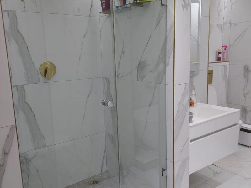 Modern bathroom featuring a glass-enclosed walk-in shower with gold-toned fixtures, marble-look wall tiles, and a sleek wall-mounted vanity unit.