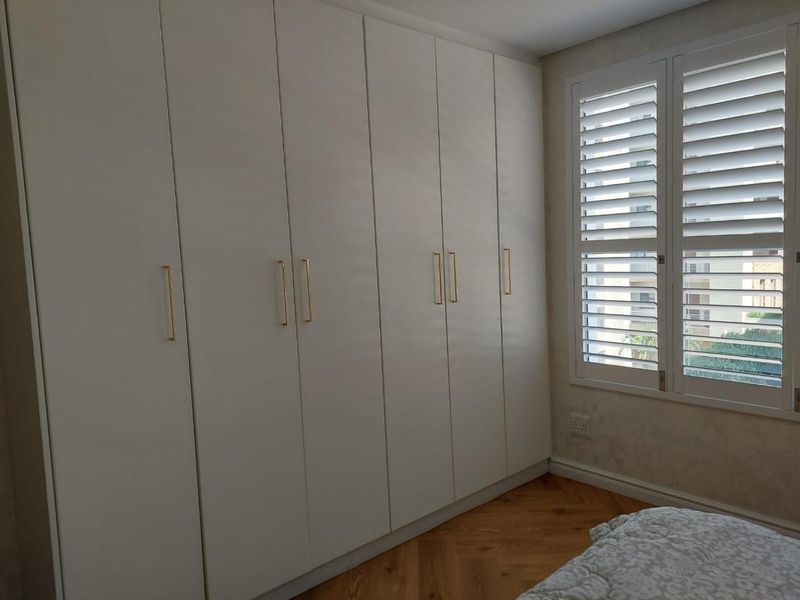 A bright bedroom featuring floor-to-ceiling built-in wardrobes with modern gold handles, herringbone timber flooring, and white plantation shutters.