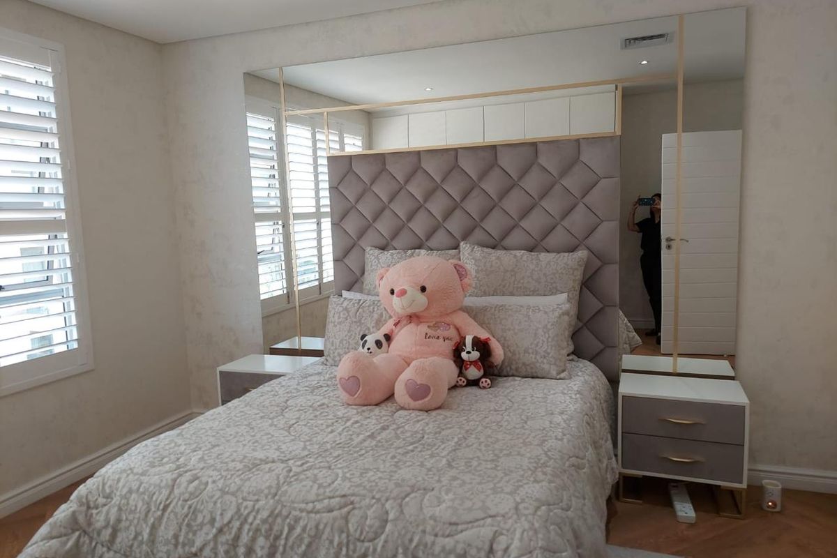 A well-appointed bedroom featuring a large tufted headboard, integrated mirrored wall panels, recessed ceiling lighting, and white plantation shutters.