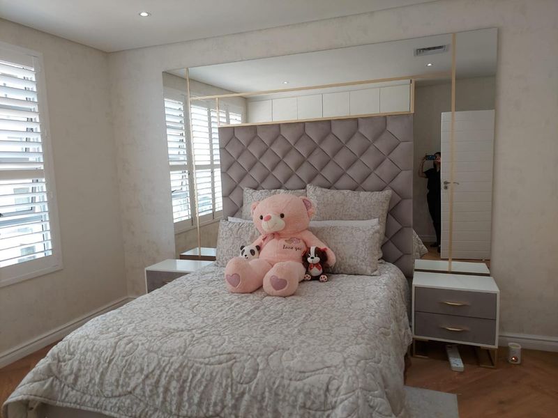 A well-appointed bedroom featuring a large tufted headboard, integrated mirrored wall panels, recessed ceiling lighting, and white plantation shutters.