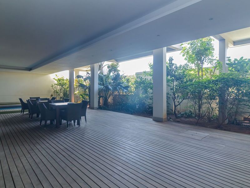 Spacious covered patio featuring timber decking, recessed lighting, and integrated garden beds, providing a seamless transition to the outdoor pool area.