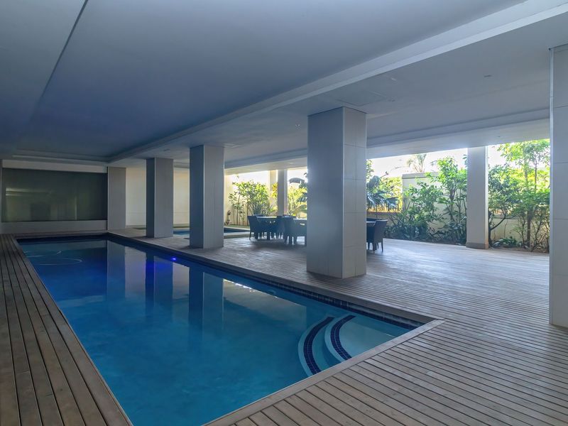A modern covered swimming pool area featuring extensive timber decking, structural columns, and integrated lighting, creating a sophisticated outdoor leisure space.