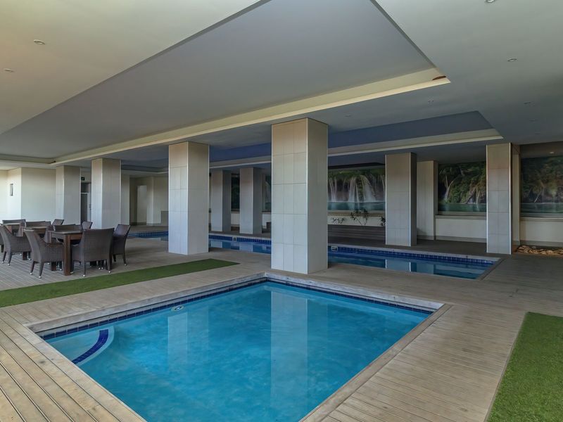 A covered indoor swimming pool area featuring tiled structural columns, recessed ceiling lighting, and wooden decking surrounding the pool.