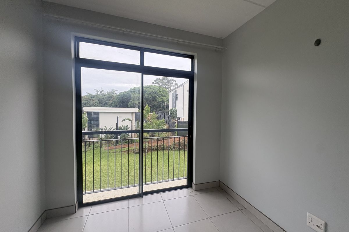 A bright, empty bedroom featuring neutral tiled flooring and a large sliding glass door that opens onto a private balcony with garden views.