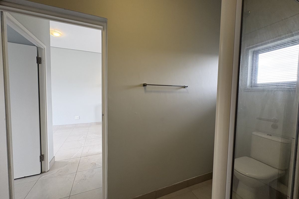 A clean, neutral bathroom featuring tiled flooring, a toilet, and a shower enclosure, with an adjacent view into an empty room.