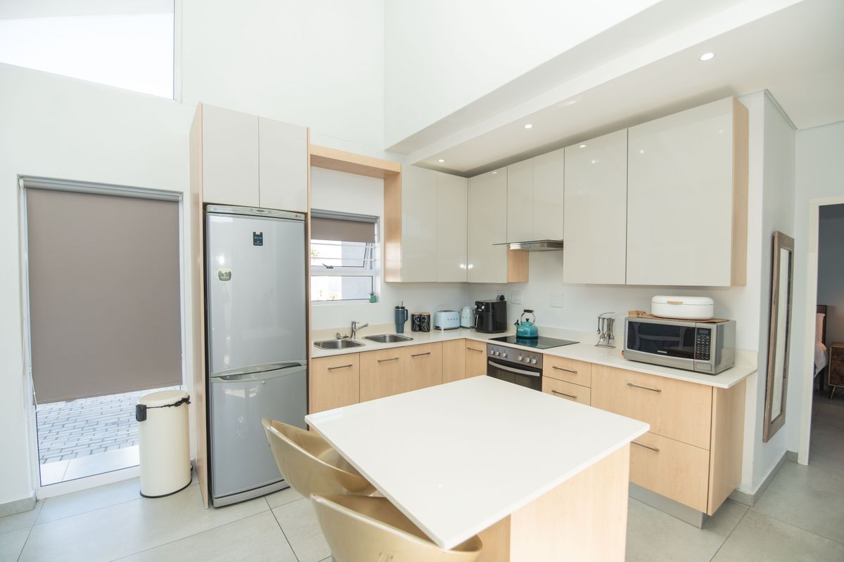 Modern kitchen featuring light wood cabinetry, stone countertops, a central island, and integrated stainless steel appliances, set within a bright, high-ceilinged open-plan space.