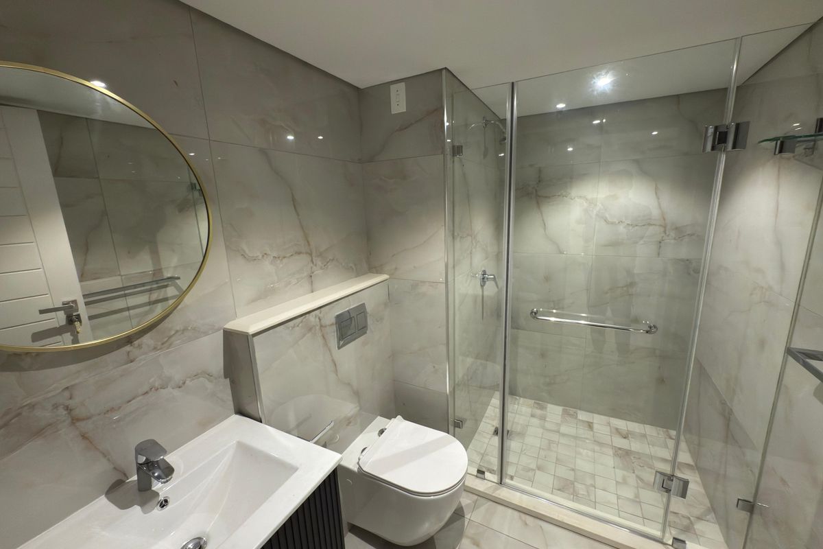 Modern bathroom featuring marble-look wall and floor tiles, a glass-enclosed walk-in shower, wall-mounted toilet, and a contemporary vanity with a circular mirror.