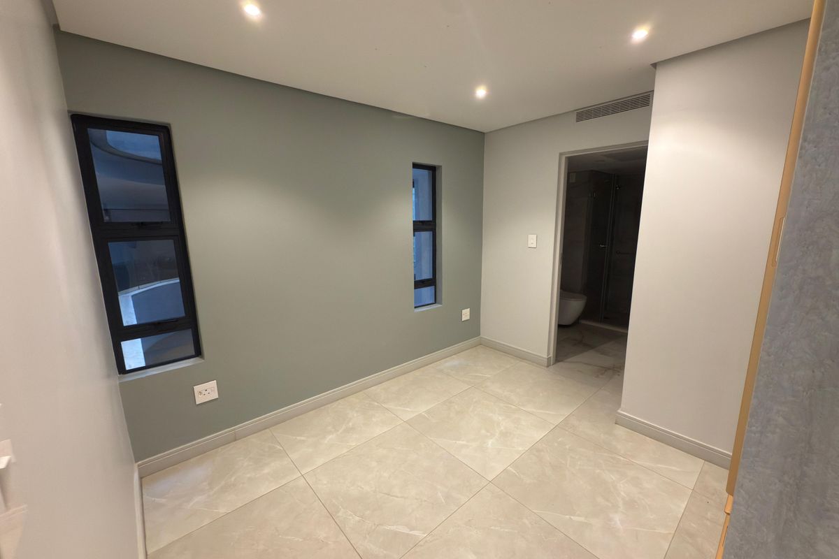 A bright, modern bedroom featuring neutral-toned large format floor tiles, recessed downlighting, and an en-suite bathroom entrance.