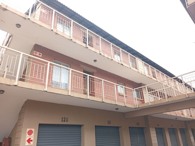 Exterior view of a multi-story apartment complex featuring brick facade, metal balcony railings, and a row of ground-level roller shutter garage doors.