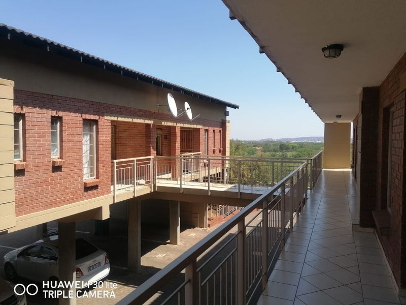 A covered balcony with tiled flooring and metal balustrades, overlooking the apartment complex grounds and surrounding landscape.