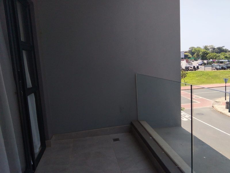 A modern balcony featuring tiled flooring and a frameless glass balustrade, offering an open view of the surrounding street and neighborhood.