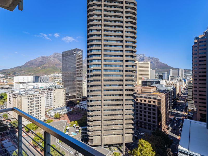 An elevated city view overlooking urban high-rise buildings and the surrounding mountain landscape, captured from a balcony with a metal railing.