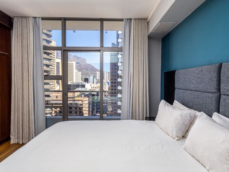 Modern bedroom featuring built-in cabinetry, hardwood flooring, and floor-to-ceiling windows offering expansive city and mountain views.