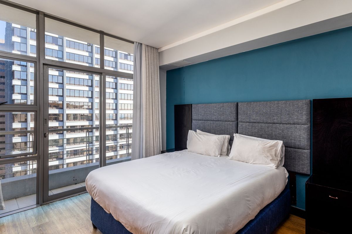 Modern bedroom featuring floor-to-ceiling windows with balcony access, a built-in headboard, and laminate flooring, offering an urban city view.