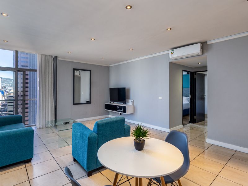 A bright open-plan living area featuring tiled floors, recessed lighting, a wall-mounted air conditioning unit, and access to a balcony with city views.