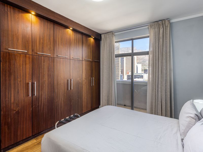 A well-maintained bedroom featuring extensive dark wood built-in cupboards with integrated lighting and a sliding door providing access to a balcony with city views.