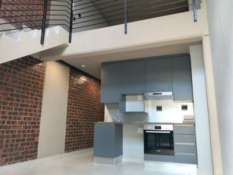 Modern open-plan kitchen featuring sleek grey cabinetry, built-in oven, extractor fan, and a striking exposed brick feature wall with an adjacent staircase.