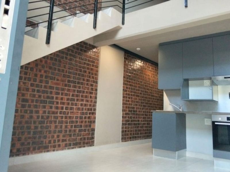 Modern open-plan kitchen featuring sleek grey cabinetry, built-in oven, and hob, set against a striking exposed brick feature wall and staircase.