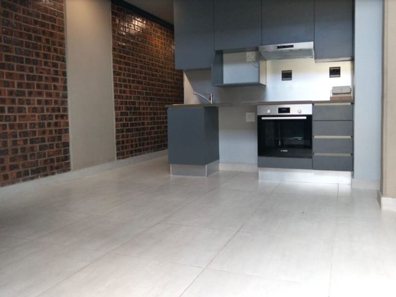 Modern open-plan kitchen featuring dark grey cabinetry, built-in oven, extractor fan, and a distinctive exposed brick feature wall with light tiled flooring.