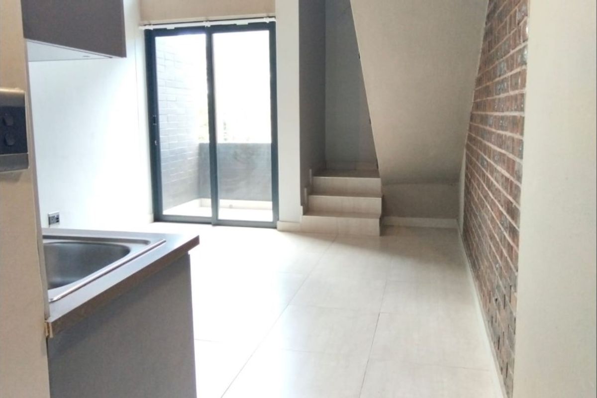 Open plan living area featuring a modern kitchen corner, exposed brick feature wall, tiled flooring, and sliding glass doors leading to a private balcony.