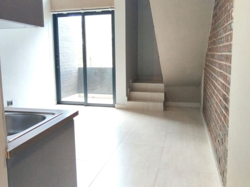 Open plan living area featuring a modern kitchen corner, exposed brick feature wall, tiled flooring, and sliding glass doors leading to a private balcony.