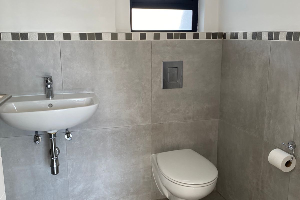 A modern bathroom featuring a wall-mounted toilet with a concealed cistern, a wall-hung basin, and neutral grey floor-to-ceiling tiling.
