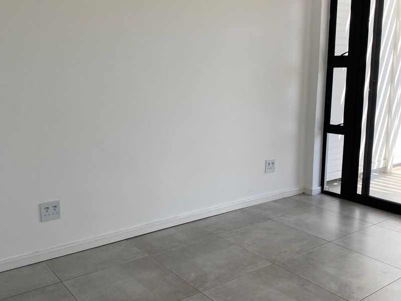 A bright, empty living area featuring modern grey floor tiles and a black-framed glass door leading to an outdoor space.