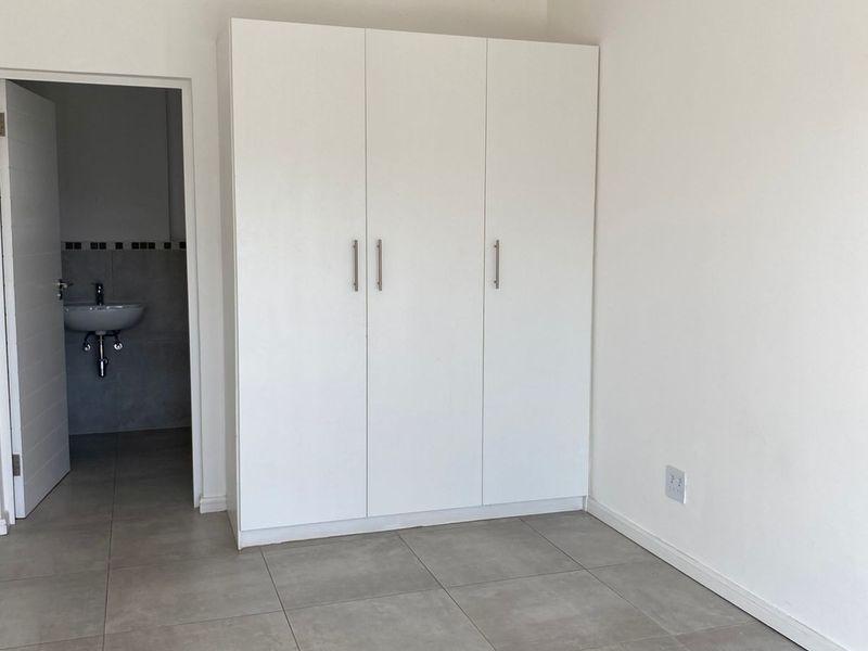 A bright bedroom featuring neutral floor tiles, built-in white cabinetry, and an adjoining en-suite bathroom with a wall-mounted basin.