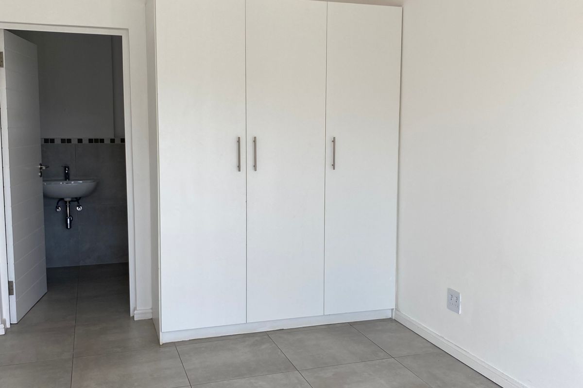 A bright bedroom featuring neutral floor tiles, built-in white cabinetry, and an adjoining en-suite bathroom with a wall-mounted basin.