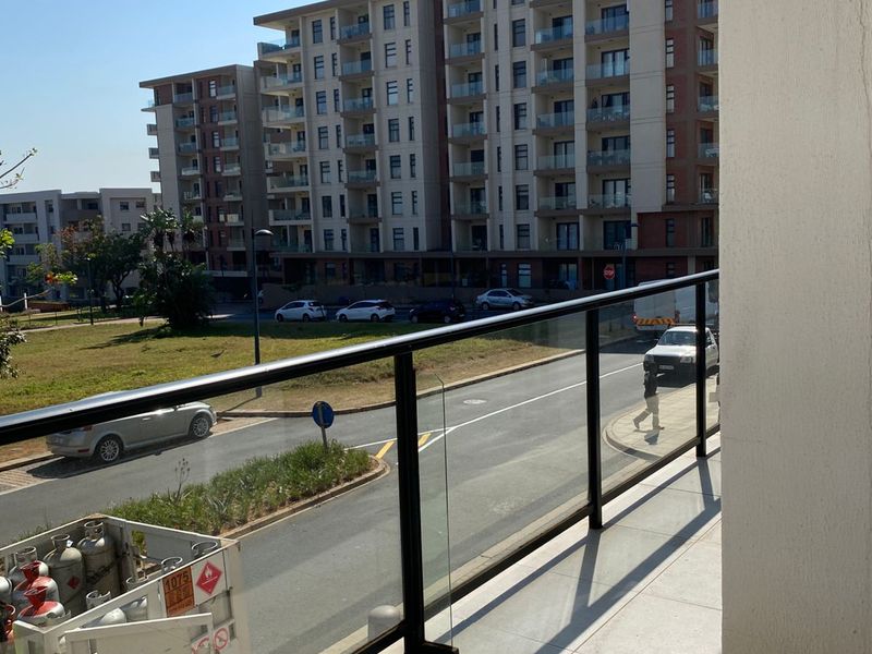 A modern apartment balcony featuring glass balustrades with metal framing, overlooking a suburban street and neighbouring residential apartment blocks.
